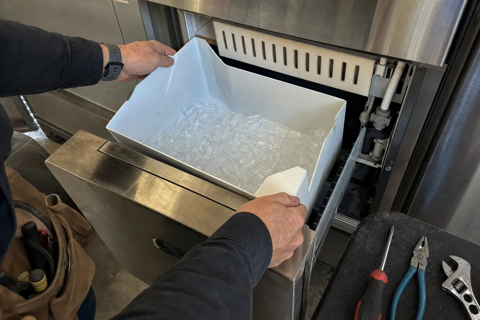 Hands lifting a full ice bin out of an undercounter ice machine, screwdriver, pliers and wrench on the mat alongside