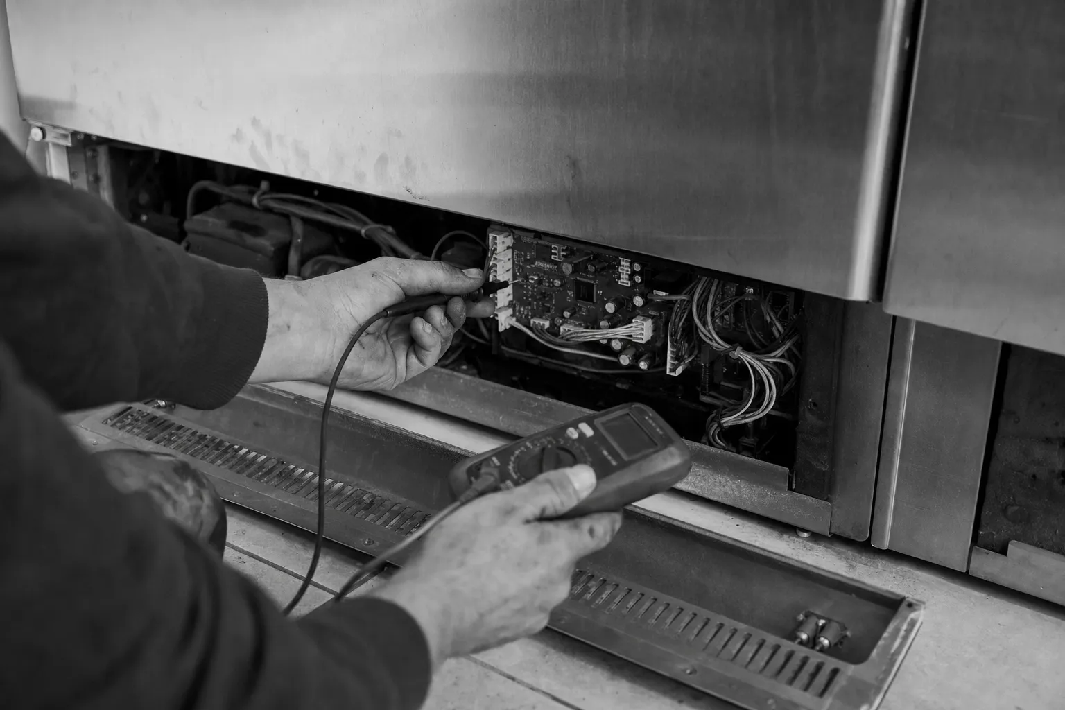 Black-and-white view of a hand probing an exposed control board behind the base panel of a stainless cabinet