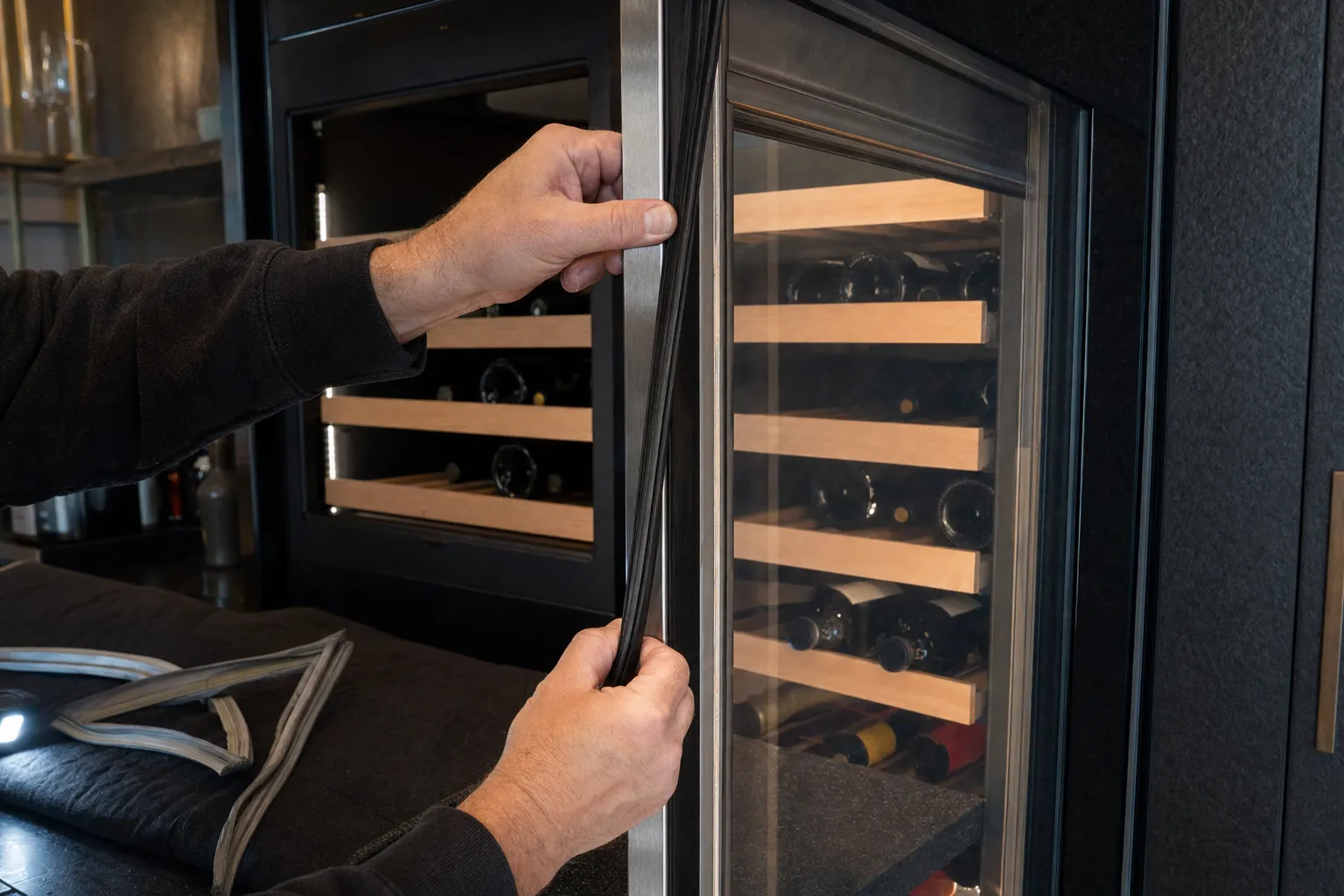 Hands fitting a new door gasket to a glass-door wine cabinet stacked with bottles on wooden racks