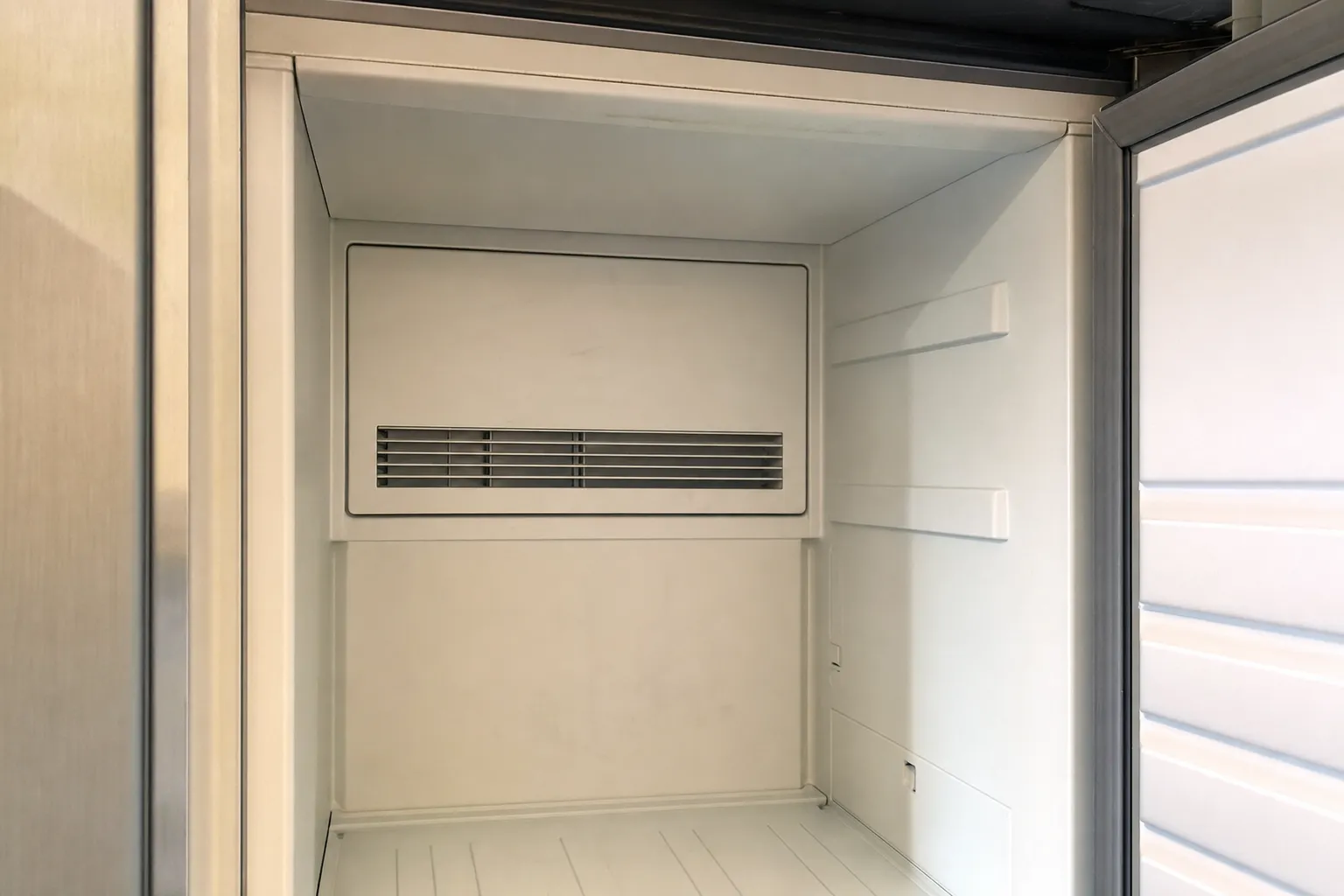 Empty white compartment of a built-in refrigeration unit with the door standing open, a louvred vent in the rear cover and no shelves or food
